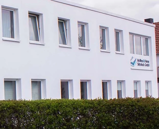 A white two-story building with multiple windows, a hedge in front, a small tree to the left, and a sign on the upper right reading "Krebs Wasser Schutz GmbH.