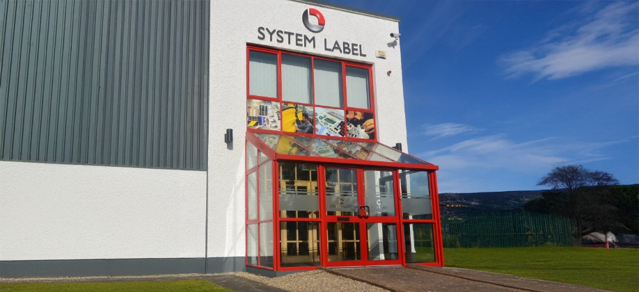 The exterior of a building with "System Label" sign above a red-framed glass entrance, set against a blue sky and green landscape.