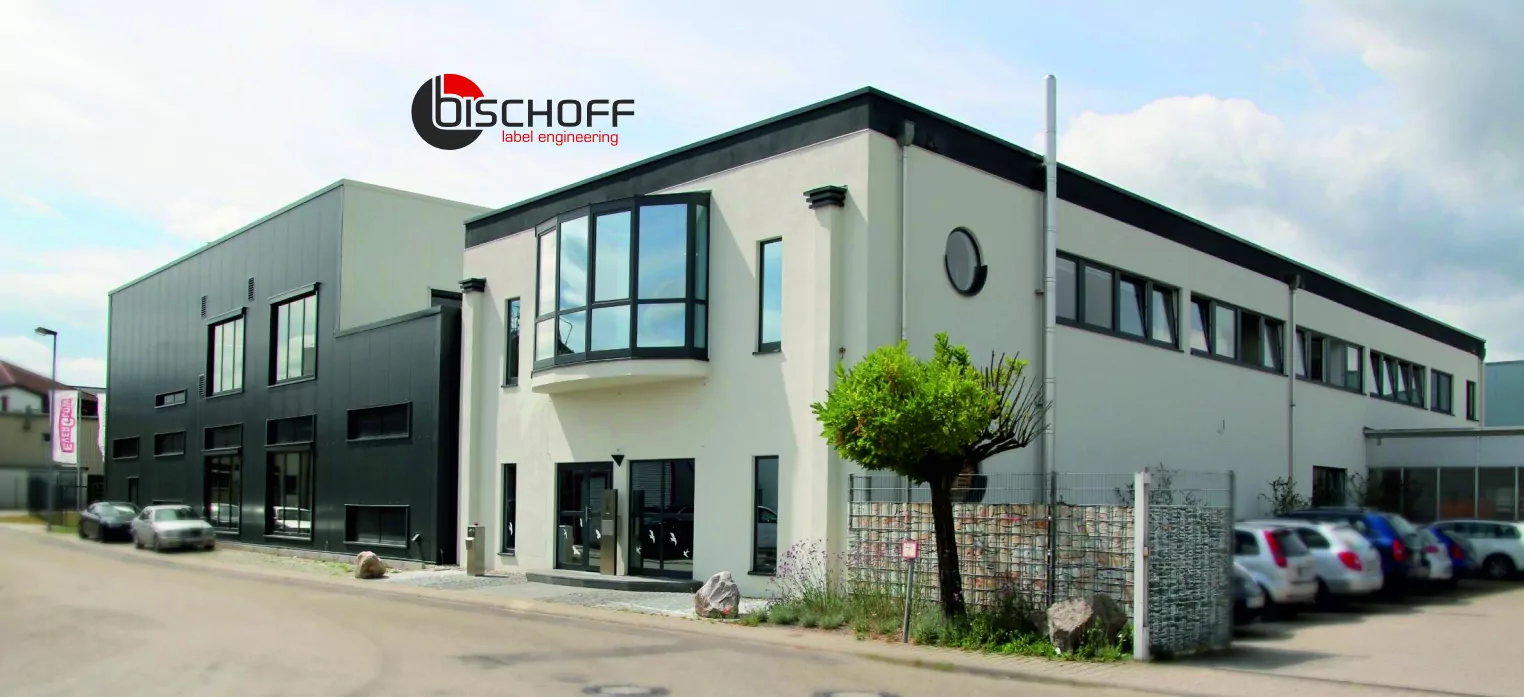 Exterior view of a modern industrial building with a "Bischoff" logo. The structure features large windows and a mix of white and gray walls. Several cars are parked nearby.