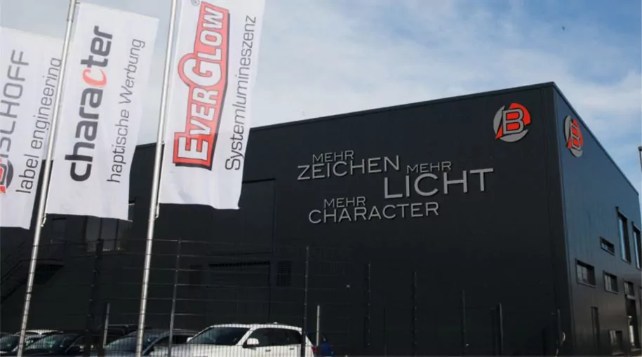 Exterior of a modern industrial building with banners. The building features logos and text, including "MEHR ZEICHEN, MEHR LICHT, MEHR CHARACTER.