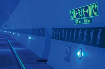 A tunnel interior illuminated by blue lights, with glowing emergency exit signs and pictograms showing directions and distances to exits.
